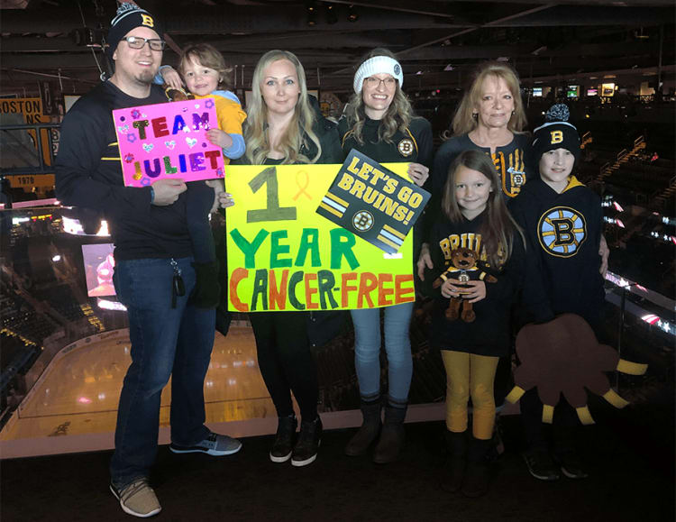 Group at a Boston Bruins hockey arena holding signs reading "TEAM JULIET" and "1 YEAR CANCER-FREE" with a yellow ribbon and "LET'S GO BRUINS!" cheering for cancer survivorship.
