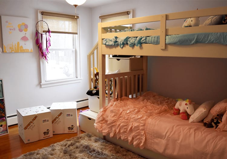 Children's bedroom with a cream-colored wooden bunk bed featuring blue bedding on the top and pink ruffled bedding on the bottom. The room has hardwood floors, a fluffy beige rug, two white storage boxes labeled "Cowpokes Juliet" and "Cowpokes Ellie," a window with beige blinds, a pink dreamcatcher, and a wall art with the text "oh the places you'll go" in yellow and pink.