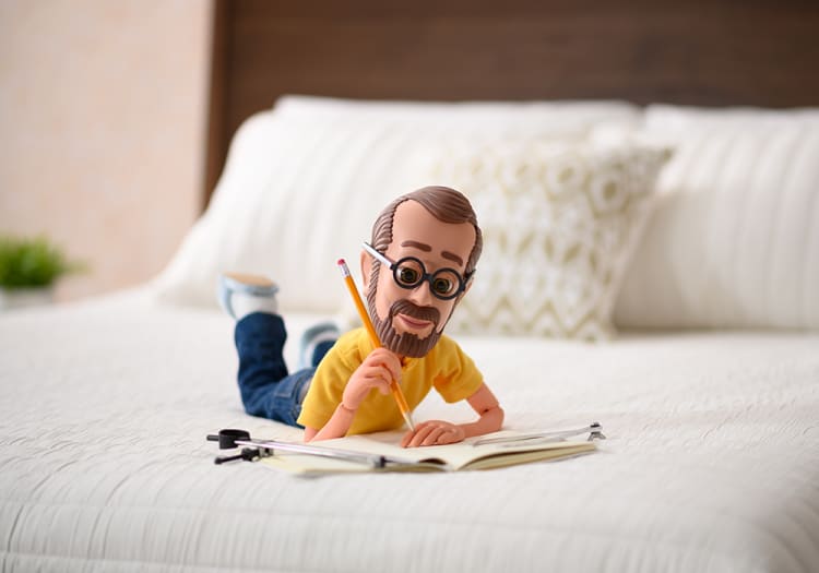 A detailed figurine of a man lying on a white bed with a yellow shirt and blue jeans, holding a pencil and writing in an open notebook.