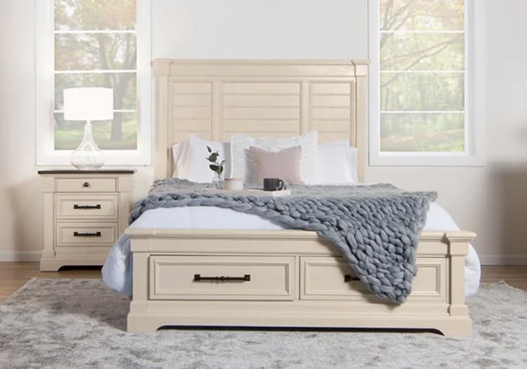 Cream-colored wooden bed frame with paneled headboard and footboard, white bedding, gray chunky knit throw, and matching nightstand with lamp.
