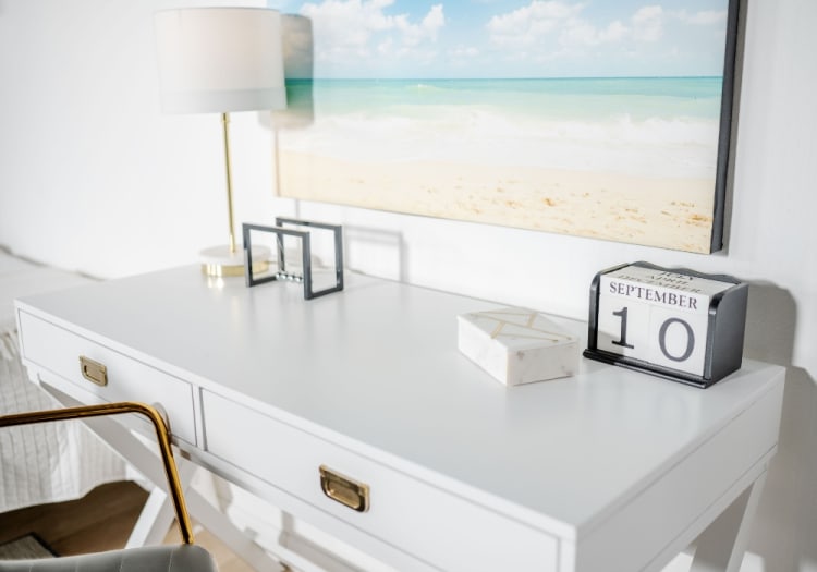 White modern desk with gold-accented lamp, marble decorative box, Newton's cradle, and a black perpetual calendar showing "September 10" under a beach-themed wall art.