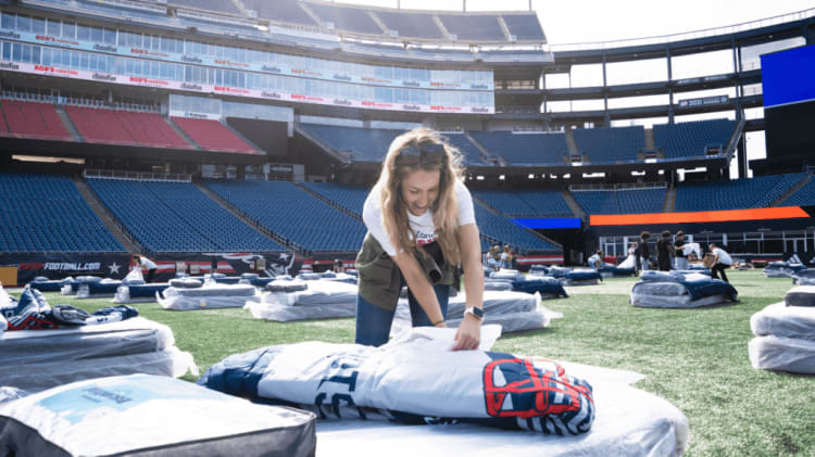 A person arranging wrapped bedding sets on a football field with empty stadium seats in the background, suggesting event setup or distribution.