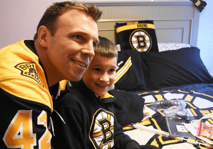 A man and child wearing black and yellow Boston Bruins hockey apparel sit on a bed with matching Bruins-themed bedding, including a pillow and blanket featuring the team logo. Various Bruins merchandise and items are scattered on the bed.