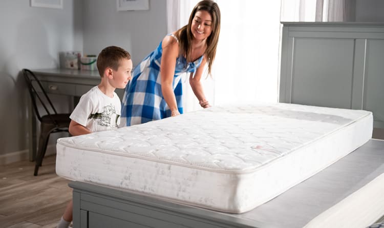 A woman and a child placing a white, quilted mattress with floral patterns on a gray wooden bed frame in a bright bedroom with light wood flooring.