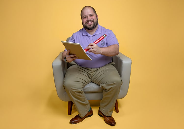 Man sitting on a gray armchair against a yellow background, wearing a lavender polo shirt with a logo, khaki pants, and brown leather shoes, holding a large red pencil and a sketchpad.