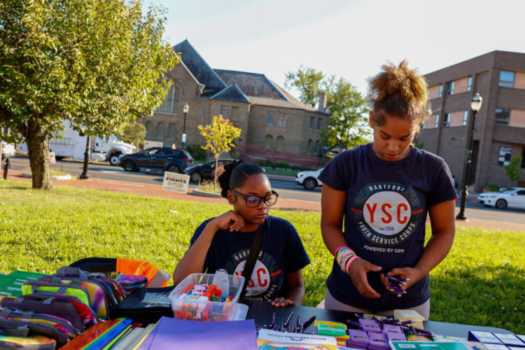 Two individuals wearing navy blue Hartford Youth Service Corps (YSC) t-shirts stand behind a table with colorful school supplies, including glue sticks, notebooks, and pencil cases, set up outdoors on a grassy area near a street.