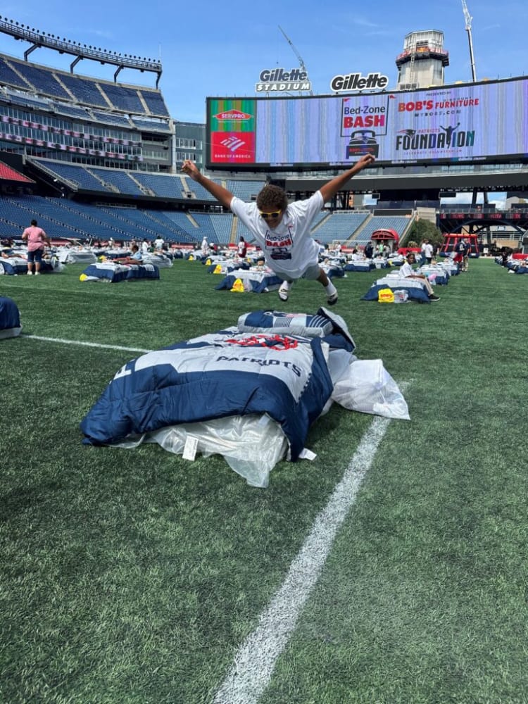 Child jumping onto a Patriots-themed sleeping bag on the field at Gillette Stadium during the Bed-Zone Bash event, with stadium seating and event signage visible.