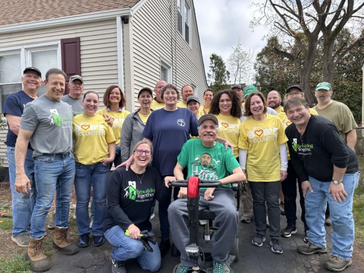 Group of volunteers wearing yellow "I ♥️ BTS VOLUNTEER" and gray or black "Rebuilding Together Manchester, CT" shirts, posing outdoors near a house.
