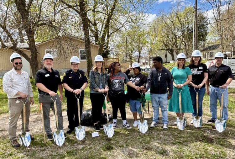 Group of people wearing white construction helmets holding shiny metal shovels, standing outdoors on a sunny day with trees and houses in the background, likely at a groundbreaking event.