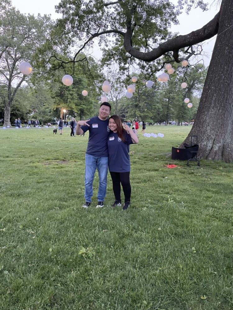 Two people wearing navy blue shirts and jeans stand on grass under a large tree with hanging white paper lanterns in a park setting during daytime.