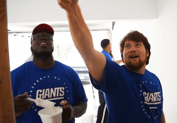 Two men wearing blue "PROPERTY OF THE GIANTS" t-shirts are painting a white wall indoors, one holding a paintbrush and bucket.
