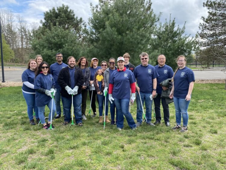 Group of people wearing blue shirts and gloves standing on grass with litter pickers, engaged in outdoor community cleanup activity near trees.
