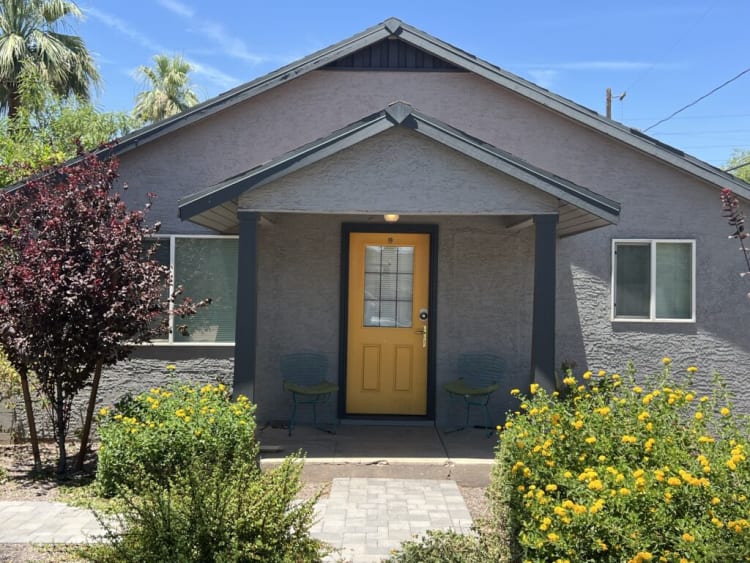 Single-story gray stucco house with a yellow front door, covered porch with two metal chairs, surrounded by green shrubs and yellow flowers under a clear blue sky.