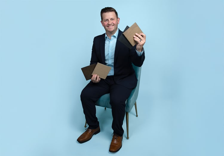 Man in a navy blue suit and light blue shirt sitting on a teal chair holding four wooden sample panels, showcasing material options.