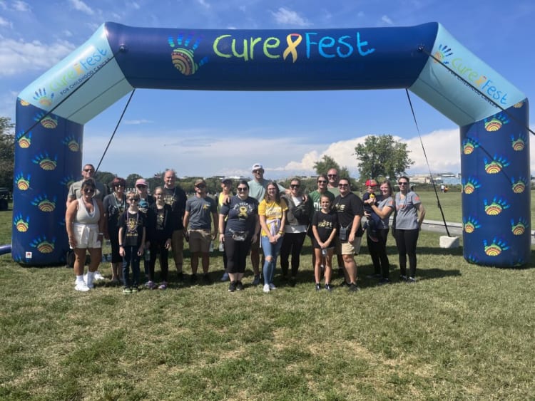 Group of people standing under a blue inflatable arch with "CureFest for Childhood Cancer" text, outdoors on grass with clear sky.