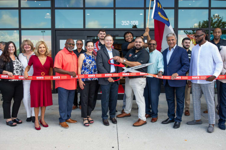 Group of people at a grand opening event holding a red ribbon with "BOB'S GRAND OPENING" text, one person cutting the ribbon with oversized scissors in front of a glass storefront.
