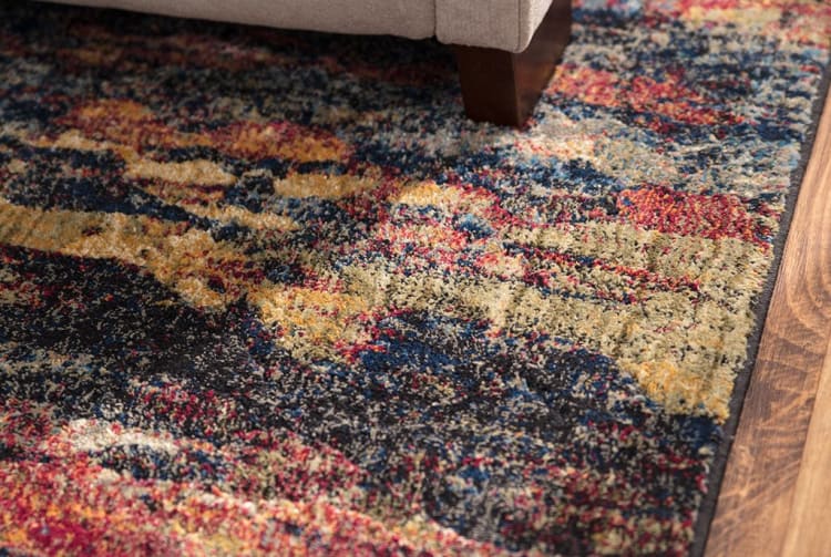 Multicolor abstract patterned area rug with a mix of dark blue, red, yellow, and beige tones, placed on wooden flooring under a beige sofa leg.