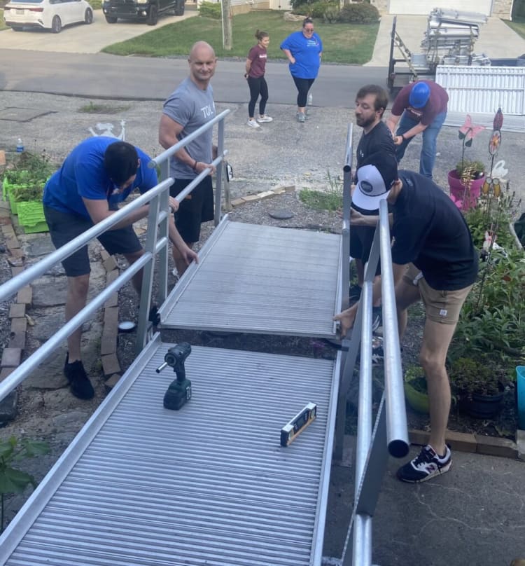 Several people installing a silver metal wheelchair ramp with handrails outdoors, using tools including a drill and level.