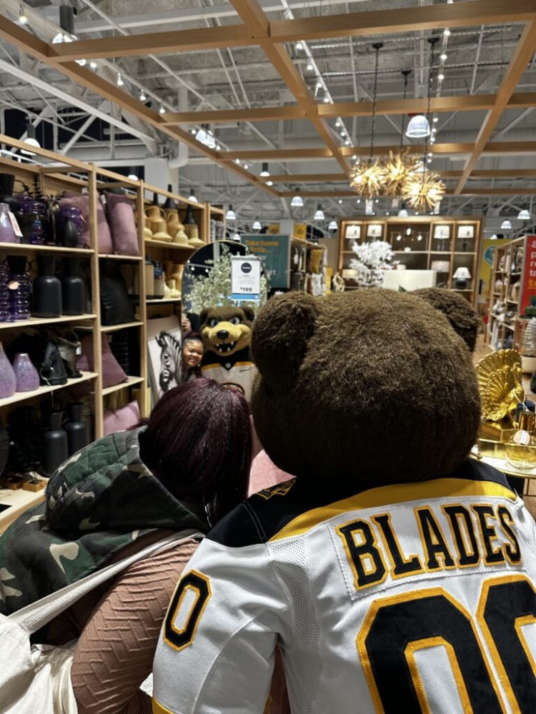 Person wearing a camouflage jacket and beige sweater standing next to a large teddy bear mascot in a white sports jersey with "BLADES" and number "00" in black and yellow, inside a well-lit home decor store with shelves of pillows, vases, and decorative items.