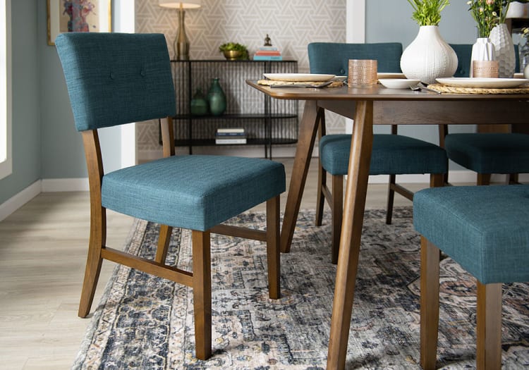 Dining room setup featuring teal upholstered chairs with wooden legs and backs, paired with a wooden table on a patterned area rug.