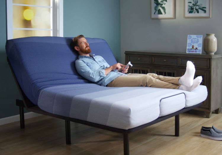 Man reclining on an adjustable blue and white striped mattress with remote control, modern bedroom setting with wooden flooring and dresser.