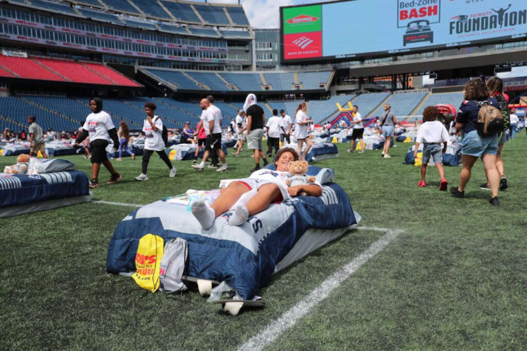 Children and adults participate in a Bed-Zone Bash event on a football field with blue and white bedding setups, featuring Bank of America and SERVPRO branding on the stadium screen.