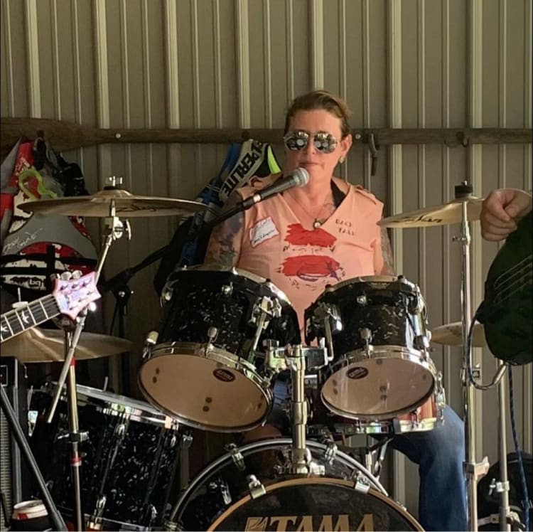 Person playing a black Tama drum set with cymbals, wearing a peach sleeveless shirt with red graphic and tattoos on arms.