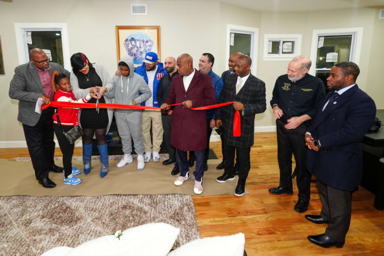 Group of people participating in a ribbon-cutting ceremony indoors, featuring a red ribbon, scissors, and diverse attendees dressed in formal and casual attire.