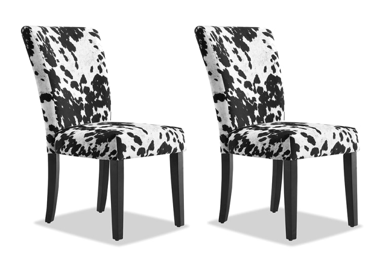 Two dining chairs with black and white cowhide-patterned upholstery, featuring cushioned seats and backs, and dark wooden legs.