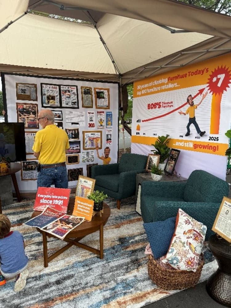 A furniture display booth with green upholstered armchairs and a wooden coffee table on a striped rug. Two banners highlight Boss Distributors Furniture's growth, featuring a chart showing 30 years of climbing furniture sales to top 100 retailers, with a red arrow rising to 2025 and text: "30 years of climbing Furniture Today's top 100 furniture retailers... BOSS Distributors Furniture" and "and still growing!" A man in a yellow shirt and jeans stands near a TV screen, with promotional signs on the table advertising "Everyday low prices since 1991" and "We sell exclusive brands you won't find anywhere else."