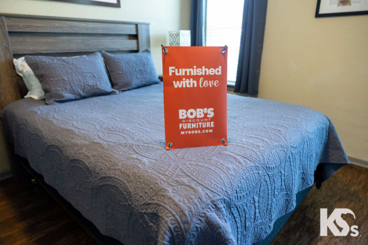 A neatly made bed with a gray quilted bedspread and matching pillows, featuring a wooden headboard. A red sign on the bed reads: "Furnished with love BOB'S DISCOUNT FURNITURE MYBOBS.COM," promoting Bob's Discount Furniture. The room has wood flooring and a window with dark curtains.