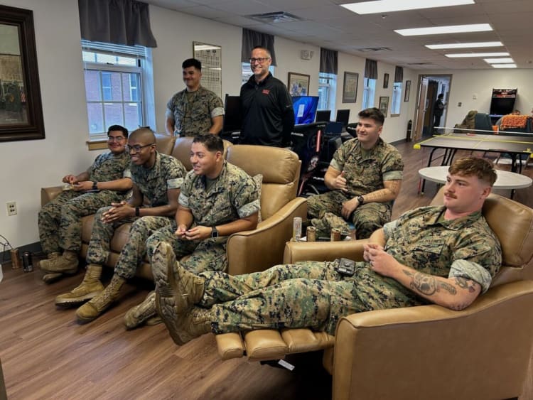 Group of military personnel in camouflage uniforms relaxing in a lounge area with leather chairs, a ping pong table, and gaming chairs in the background.