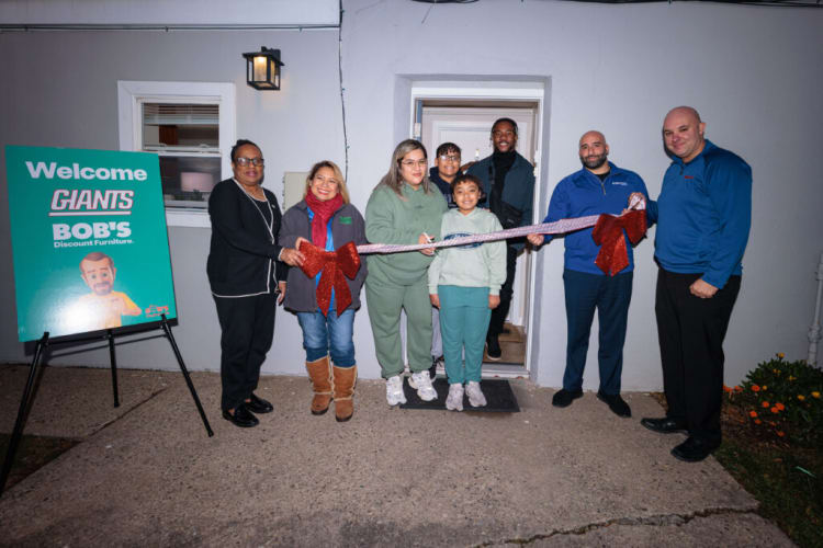 Group of people standing outside a building holding a decorative ribbon with red bows for a ribbon-cutting event. A teal sign on the left reads: "Welcome GIANTS BOB'S Discount Furniture." The scene suggests a community or business opening celebration.