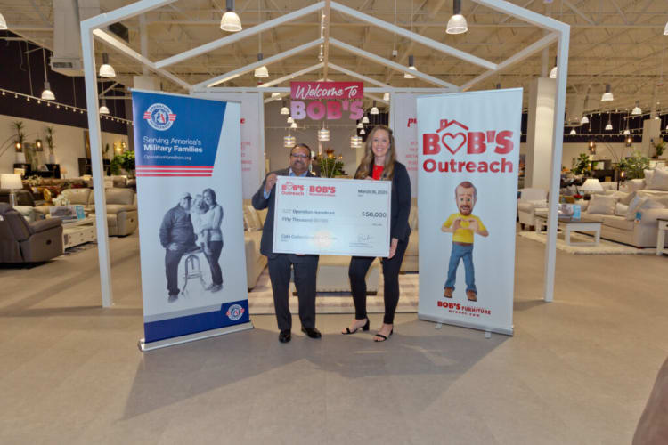 Two people standing inside a furniture store holding a large check for $50,000 dated March 16, 2023, from Bob's Outreach to Operation Homefront. Two vertical banners flank them: one reads "Serving America's Military Families" with a photo of a couple, the other shows a cartoon figure with text "Bob's Outreach" and "Bob's Furniture." A sign above says "Welcome to Bob's." The setting highlights a charitable donation event supporting military families at Bob's Furniture store.