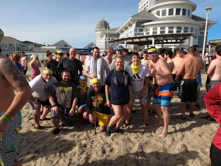 Group of people wearing casual beachwear and colorful bandanas gathered on sandy beach near large white building, sunny day.