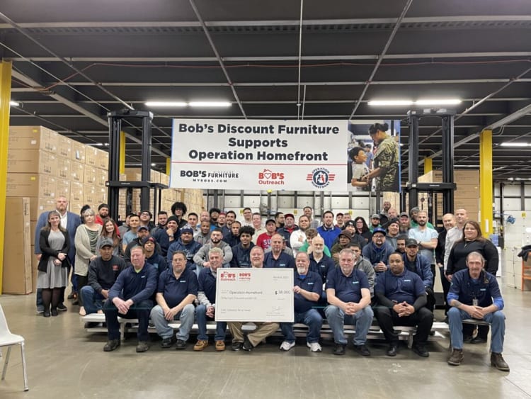 Group of employees posing in a warehouse under a banner that reads "Bob’s Discount Furniture Supports Operation Homefront" holding a large check for $38,000 from Bob’s Outreach.