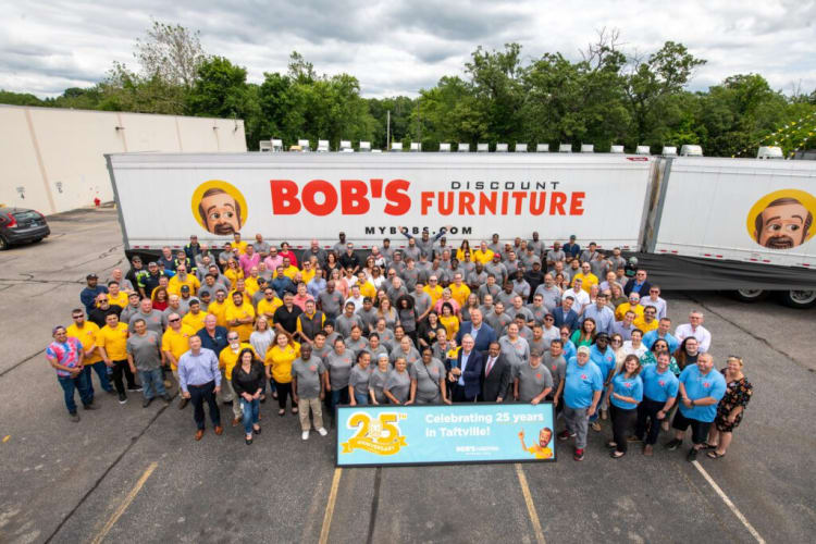 Large group of people wearing yellow, gray, blue, and casual attire standing in front of two Bob's Discount Furniture trucks. Banner reads "25th Anniversary Celebrating 25 years in Taftville! BOB'S furniture."