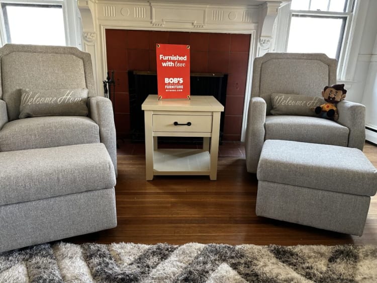 Two gray upholstered armchairs with matching ottomans and "Welcome Home" pillows flank a beige wooden side table in front of a fireplace. The side table holds a red sign that reads "Furnished with love by BOB'S DISCOUNT FURNITURE MYBOBS.COM." The room features hardwood floors and a patterned area rug.