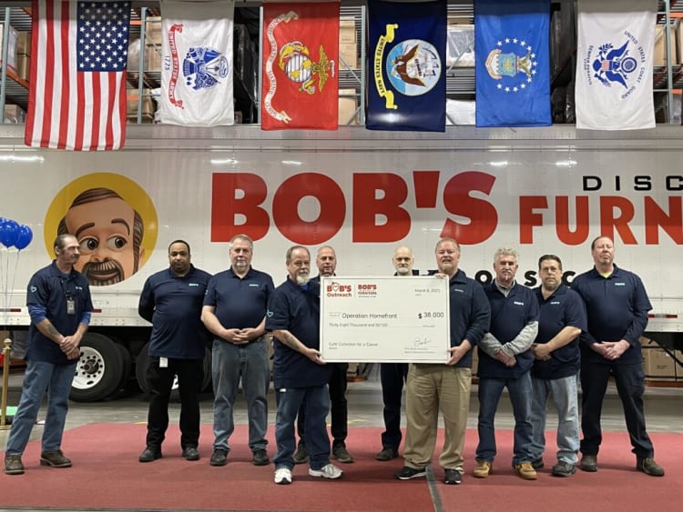 Group of employees in navy blue shirts holding a large check for $38,000 from Bob's Furniture Outreach, supporting Operation Homefront, with American and military flags displayed above.