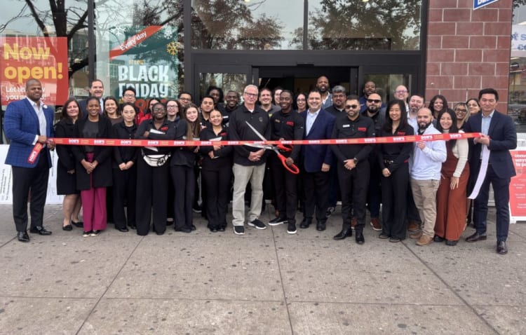 Group of people standing outside a store for a grand opening ribbon-cutting ceremony, holding a red ribbon with "BOB'S GRAND OPENING" text, with signage in the background reading "Now open!" and "Every day is like Black Friday."