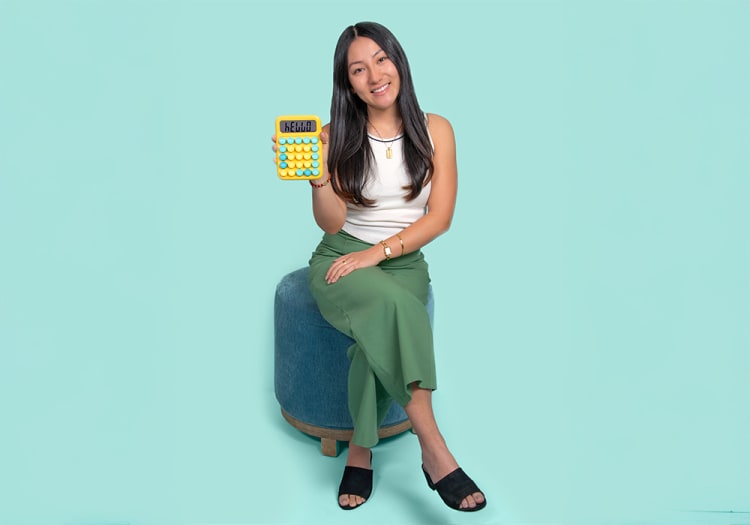 Woman sitting on a blue ottoman holding a yellow calculator displaying the word "HELLO," wearing a white tank top, green skirt, and black sandals.