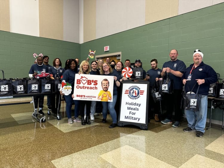 Group of people holding a sign that reads "BOB'S Outreach BOB'S Furniture MYBOBS.COM" standing next to a board with "OPERATION HOMEFRONT Holiday Meals For Military" text, with black gift bags on tables and in hands, indicating a holiday meal outreach event for military families.