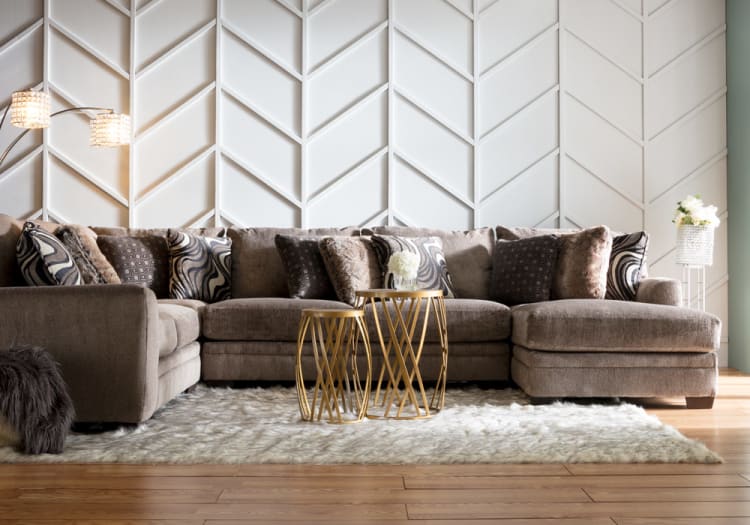 Large modern living room with a plush brown sectional sofa featuring patterned and fur-textured throw pillows, gold metal nesting coffee tables, a white textured accent wall, and a soft white shag rug on wooden flooring.