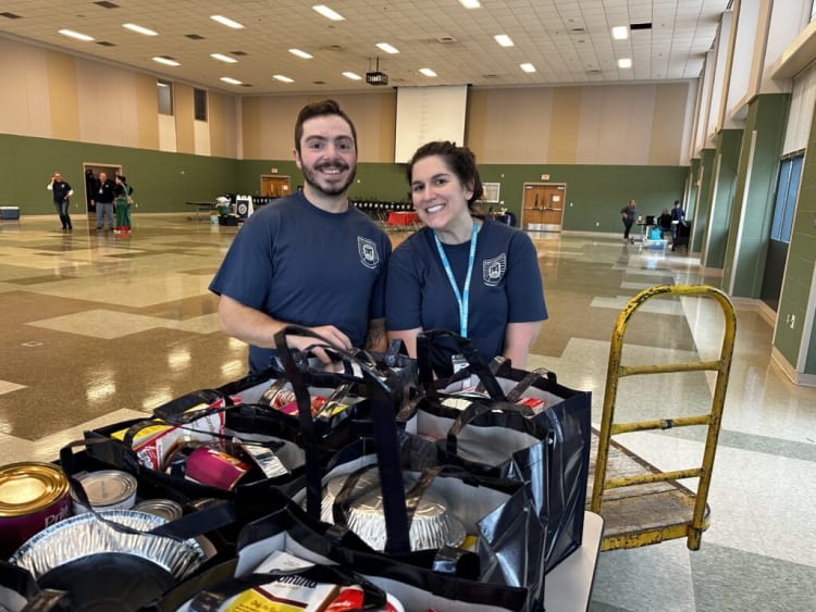 Two people in navy blue shirts stand behind a table filled with black reusable bags containing canned goods and food items in a large, well-lit community center.