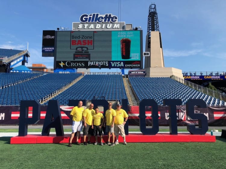 Group of five people in yellow shirts standing on a football field in front of large "PATRIOTS" letters at Gillette Stadium with a digital scoreboard displaying "Bed-Zone BASH" and sponsor logos.