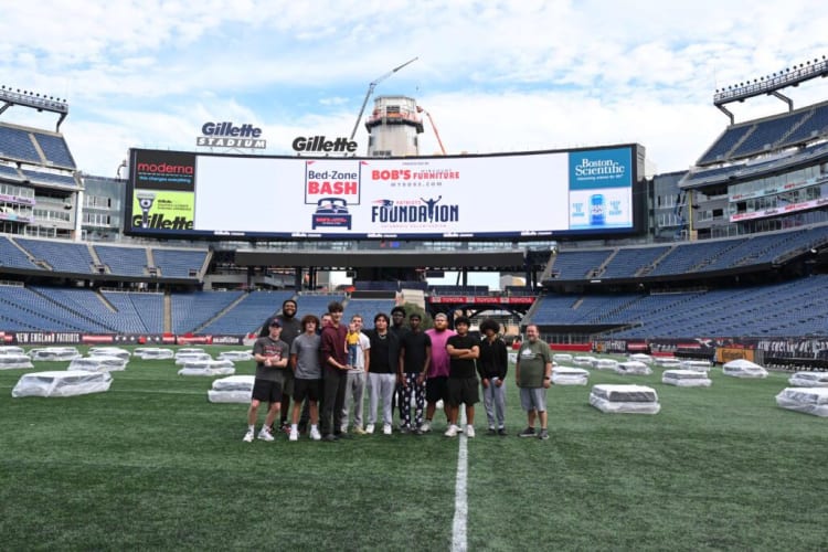 Group of young men standing on the field at Gillette Stadium with covered equipment in the background and a large digital scoreboard displaying sponsors including Moderna, Bob’s Furniture, and Boston Scientific.