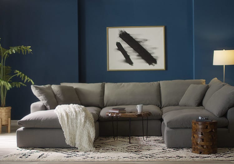 Gray sectional sofa with plush cushions and a white textured throw blanket, set against a dark blue wall with abstract black and white artwork, wooden side table, and floor lamp.