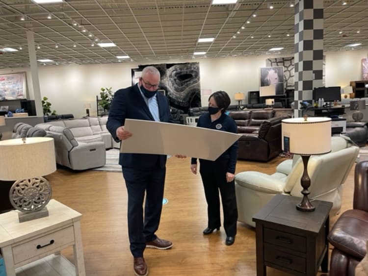 Two people in a furniture showroom examining a large beige sample board, surrounded by modern sofas, armchairs, and wooden side tables with lamps.