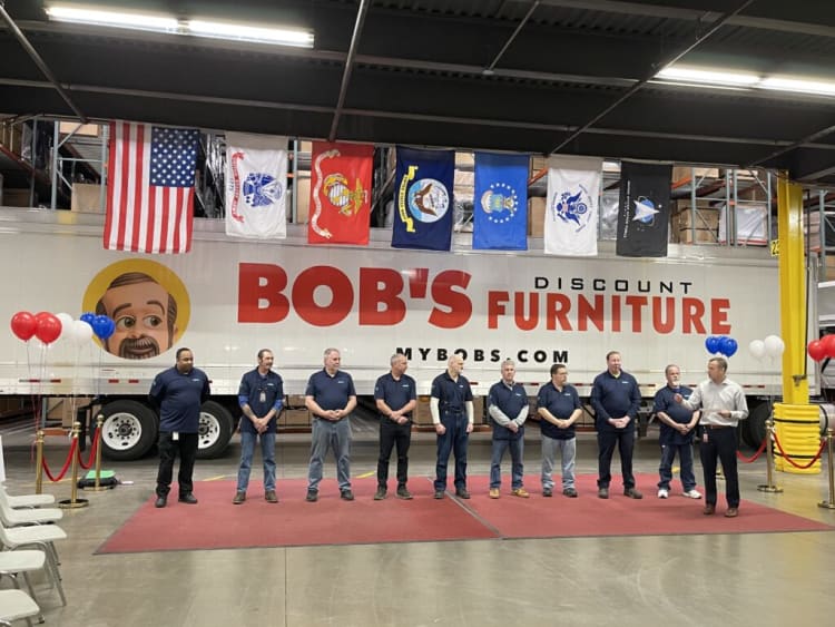 Group of employees standing in front of a large truck trailer with "BOB'S DISCOUNT FURNITURE" and "MYBOBS.COM" branding, under hanging flags including the American flag.