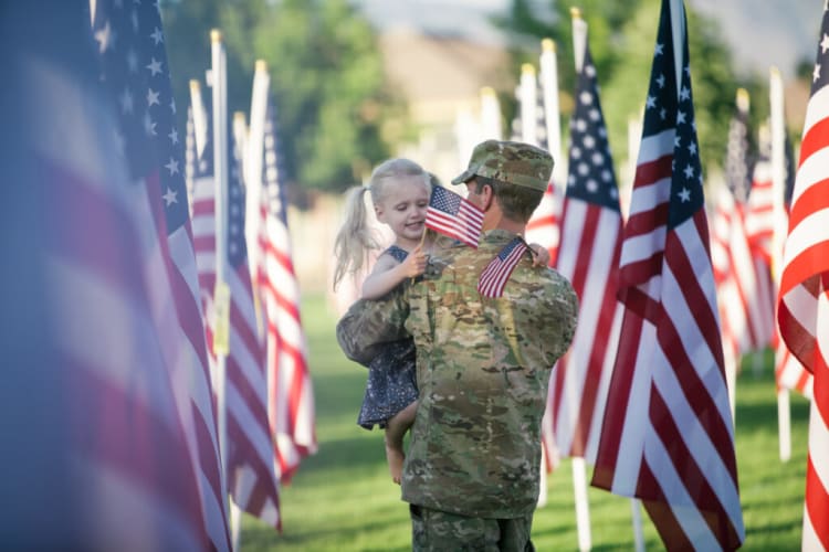 A soldier in camouflage uniform holding a child while standing among multiple American flags outdoors, symbolizing patriotism and military family.